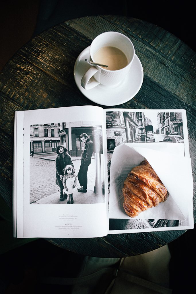 Photo of a warm croissant sitting on a black and white magazine with a creamy coffee, at a coffee shop table.