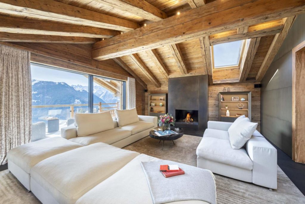 Inside of a traditional wooden chalet with a firepit, white sofas and a beautiful view of the mountains.