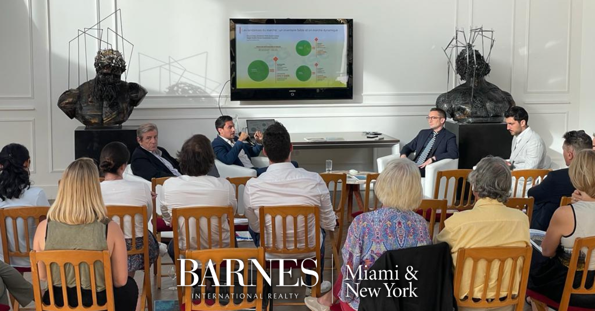Люди, сидящие на стульях, слушают конференцию, а директор BARNES в Нью-Йорке и Майами представляет PowerPoint по телевизору.
