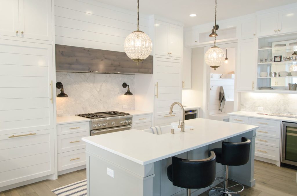 Luxury kitchen, well decorated in a white, beige palette. Two round chandeliers are hanging from the ceiling, an island countertop with two stools is in the foreground.