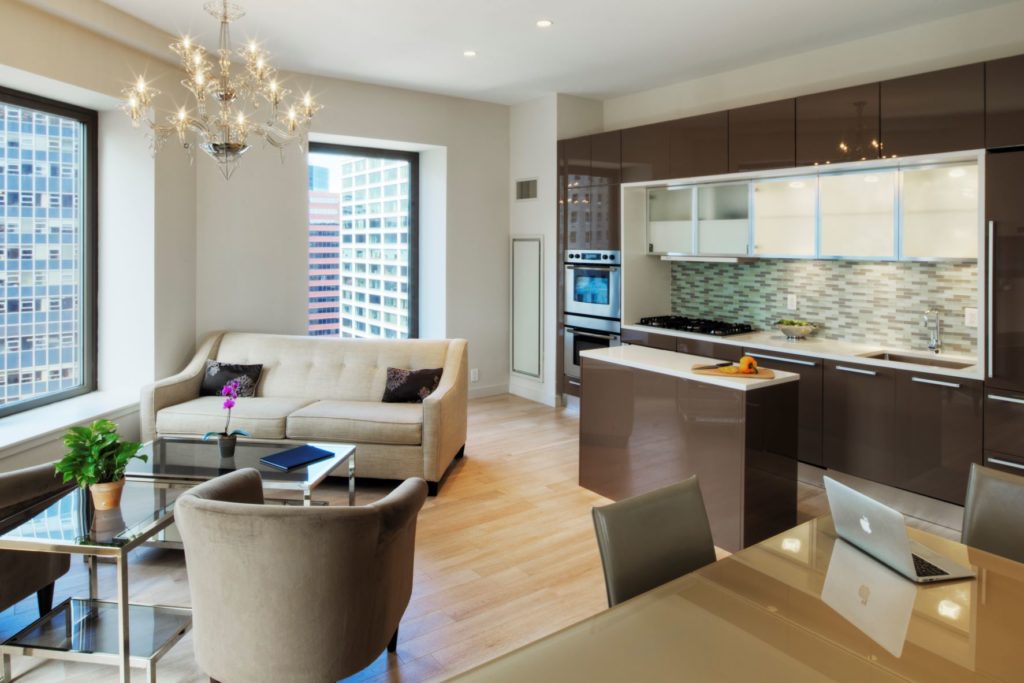 Vue de l'intérieur d'un appartement dans un immeuble de luxe, montrant la cuisine et le salon avec des fenêtres surdimensionnées et des vues urbaines.