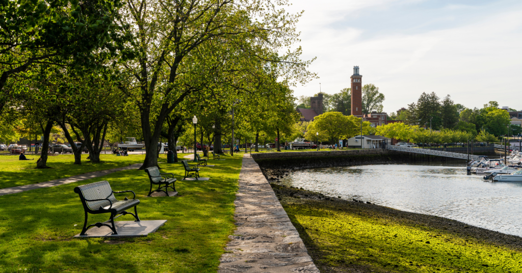 Park with benches on the water with sunshine, and a lighthouse in the background.